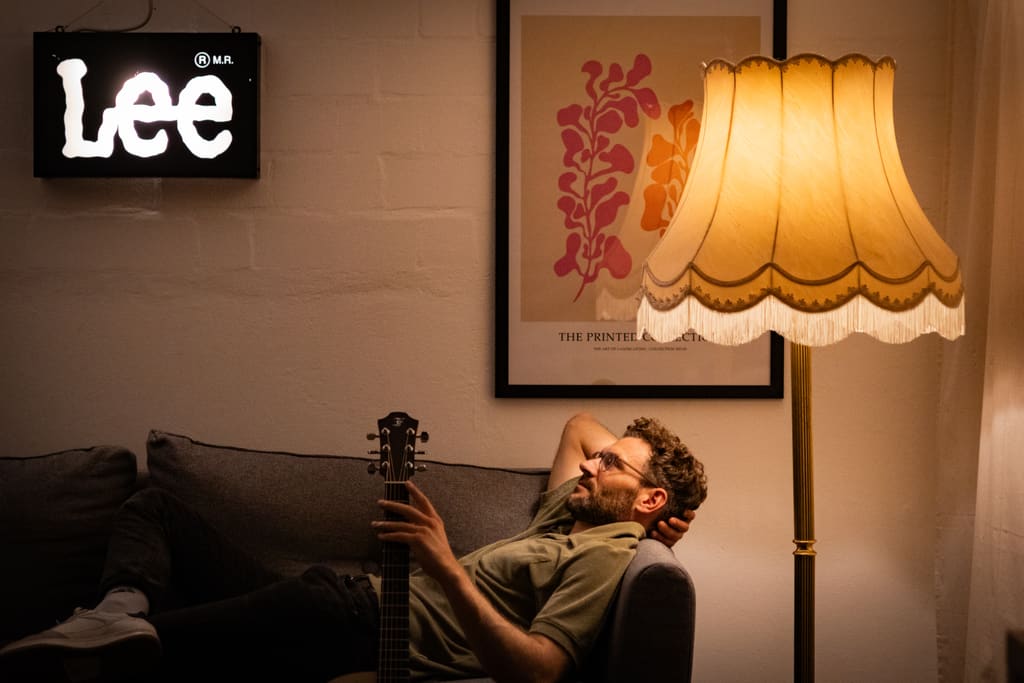 Jochen liegt mit Gitarre auf dem Sofa unter der leuchtenden Fransen-Stehlampe.